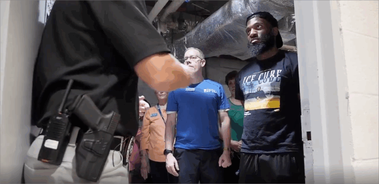 A person in a black shirt and light pants with a holstered gun and a radio on their hip stands facing a group of people. The group includes a man in a blue shirt with "FIT" and "ICE CUBE" on it, a woman in an orange shirt, and a man in a black "ICE CUBE" t-shirt with a beard and a backwards baseball cap. They are standing in a room with exposed ductwork on the ceiling.