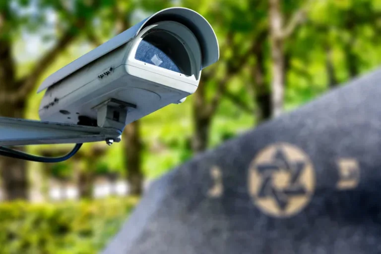 A security camera is mounted on a pole, overlooking a gravestone with a Star of David symbol.