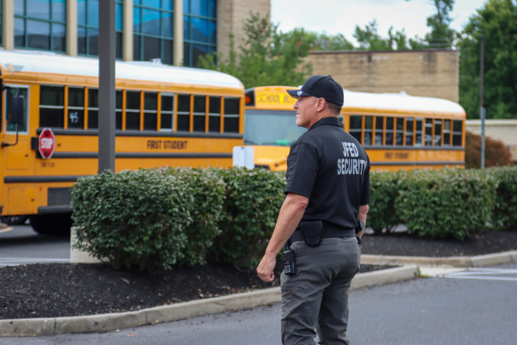 A security guard wearing a black shirt with "JFED SECURITY" on the back stands in front of yellow school buses.