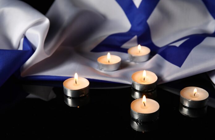 Six lit tea light candles arranged on a reflective black surface in front of a draped Israeli flag.