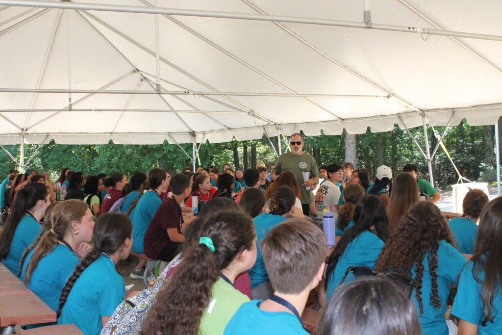 A man in sunglasses and a green t-shirt speaks to a group of children sitting under a white tent.