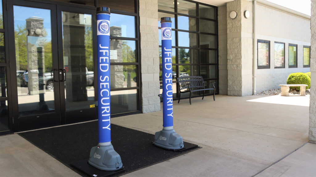Two blue security bollards with "JFED SECURITY" text and a logo stand in front of a building entrance with glass doors.