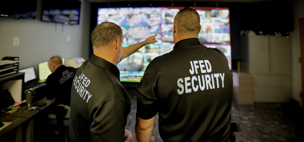 Two security guards in black shirts with "JFED SECURITY" printed on the back stand in front of a large screen displaying multiple security camera feeds. One guard is pointing at the screen.
