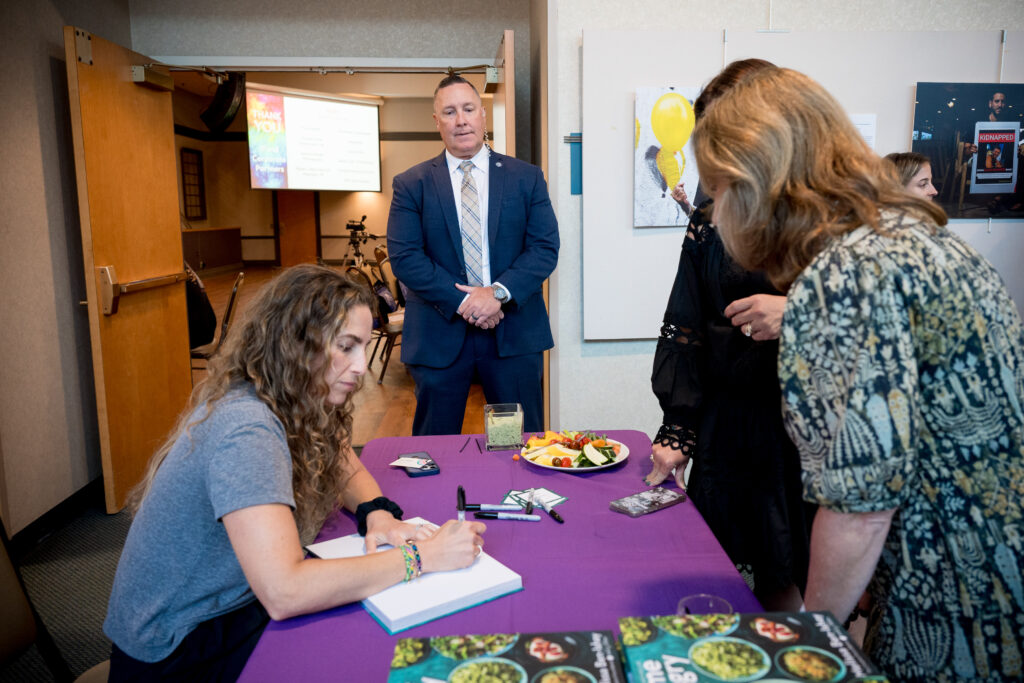 A woman with long, curly brown hair signs a book at a purple table, while a man in a suit stands behind her. Two other women are partially visible on the right, and a "THANK YOU" slide is projected on a screen in the background.