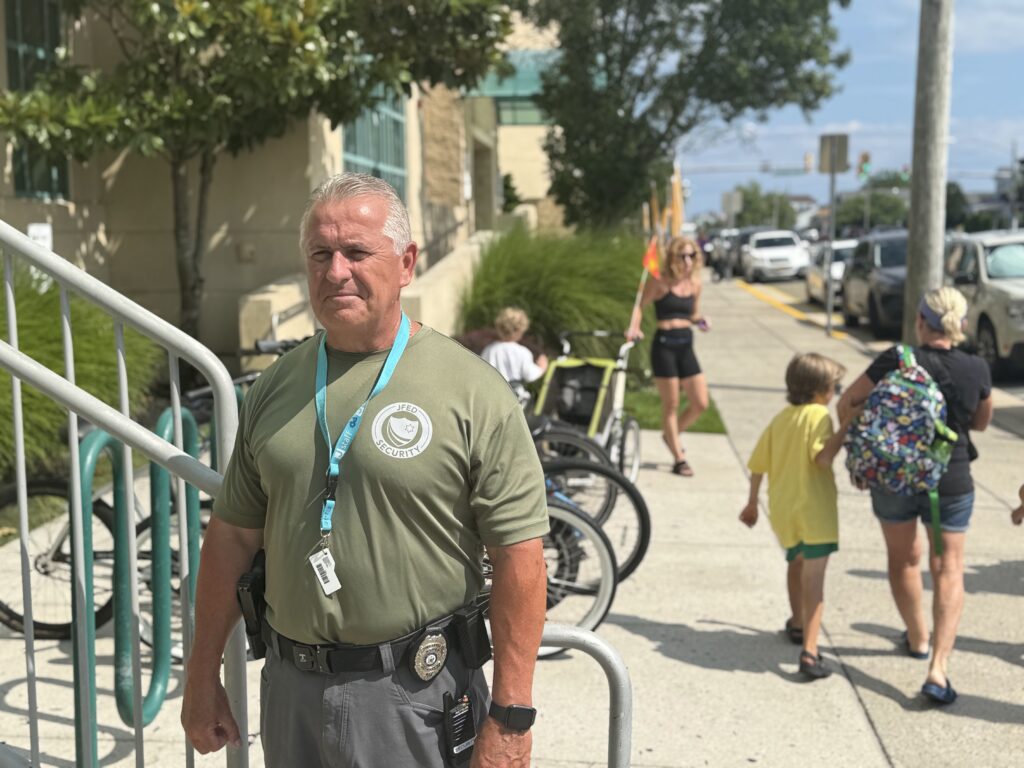 A security guard in an olive green shirt with a "JFED SECURITY" logo stands in front of a building. He is wearing a badge, a holster with a firearm, and a radio. In the background, people walk along a sidewalk with bicycles and cars.