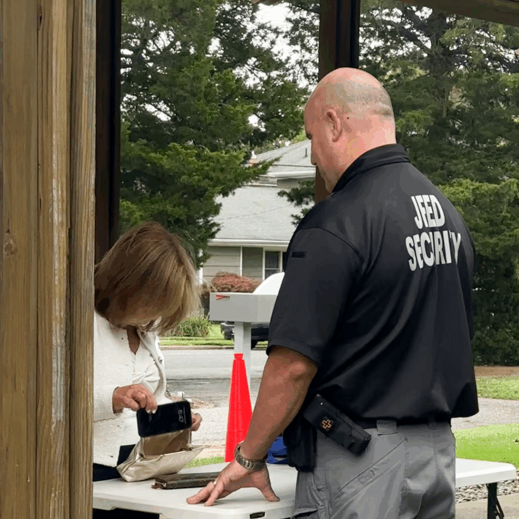 A security guard in a black shirt with "JFED SECURITY" on the back stands next to a woman in a white jacket who is putting a phone into her purse on a white table.