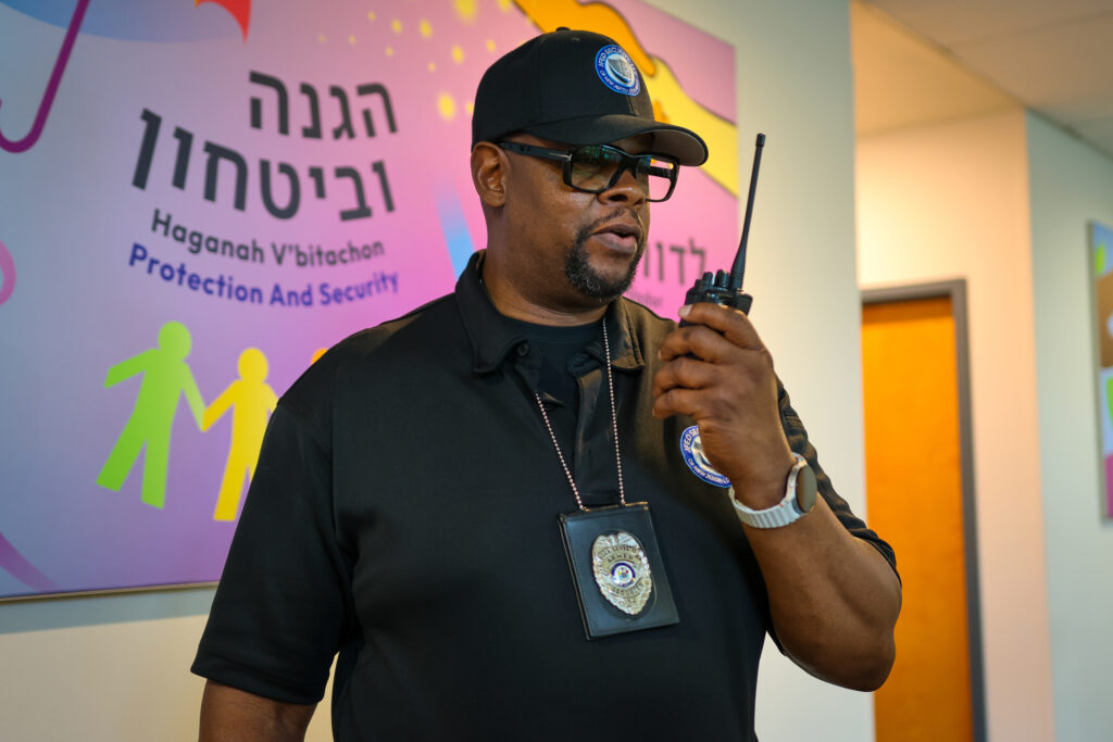 A Black security guard wearing a black polo shirt, black baseball cap, and glasses speaks into a black walkie-talkie. He has a badge clipped to his shirt. Behind him is a wall with text in Hebrew and English that reads "Haganah V'bitachon Protection And Security" and colorful graphics of people.