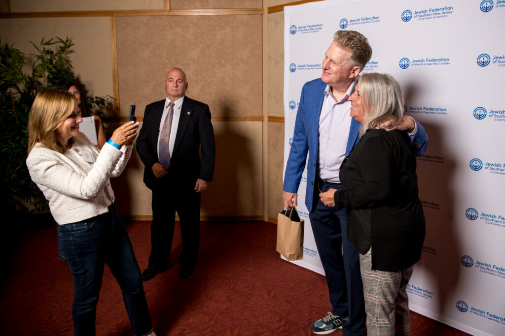 A woman in a white blazer takes a photo with her phone of a man in a blue suit and a woman in a black top, with a man in a dark suit standing behind them. A backdrop with the "Jewish Federation of Southern New Jersey & Our Family of Agencies" logo is visible.