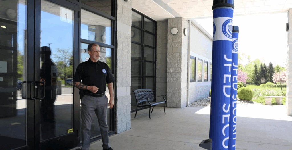 A security guard in a black polo shirt and grey pants stands outside a building with glass doors, next to a blue banner that says "JFED SECURITY.