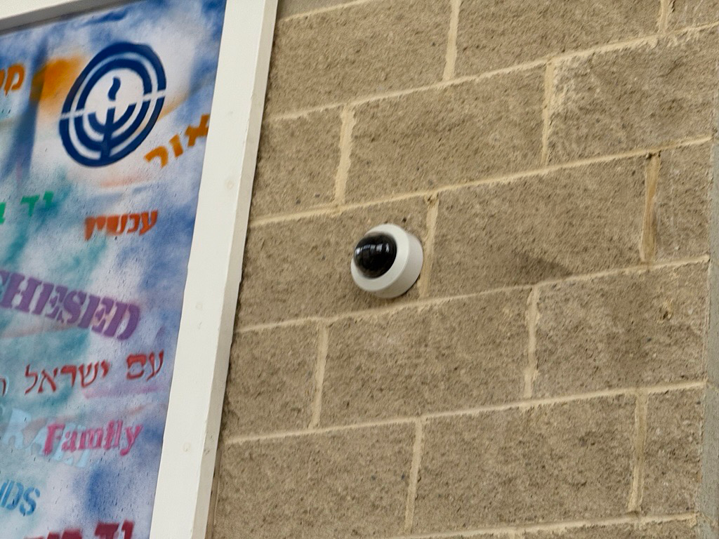 A white dome security camera is mounted on a tan brick wall next to a poster with Hebrew text and a blue menorah symbol.